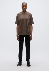 Καφέ μεγάλης διάστασης μπλούζα με στρογγυλή λαιμόκοψη και λεπτομέρεια λογοτύπου στο στήθος, σετ με μαύρο παντελόνι με ρίγες στο πλάι και μαύρα αθλητικά παπούτσια.