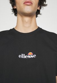 Jeune personne portant un t-shirt noir avec un petit logo ellesse blanc, rouge et orange brodé sur la poitrine.