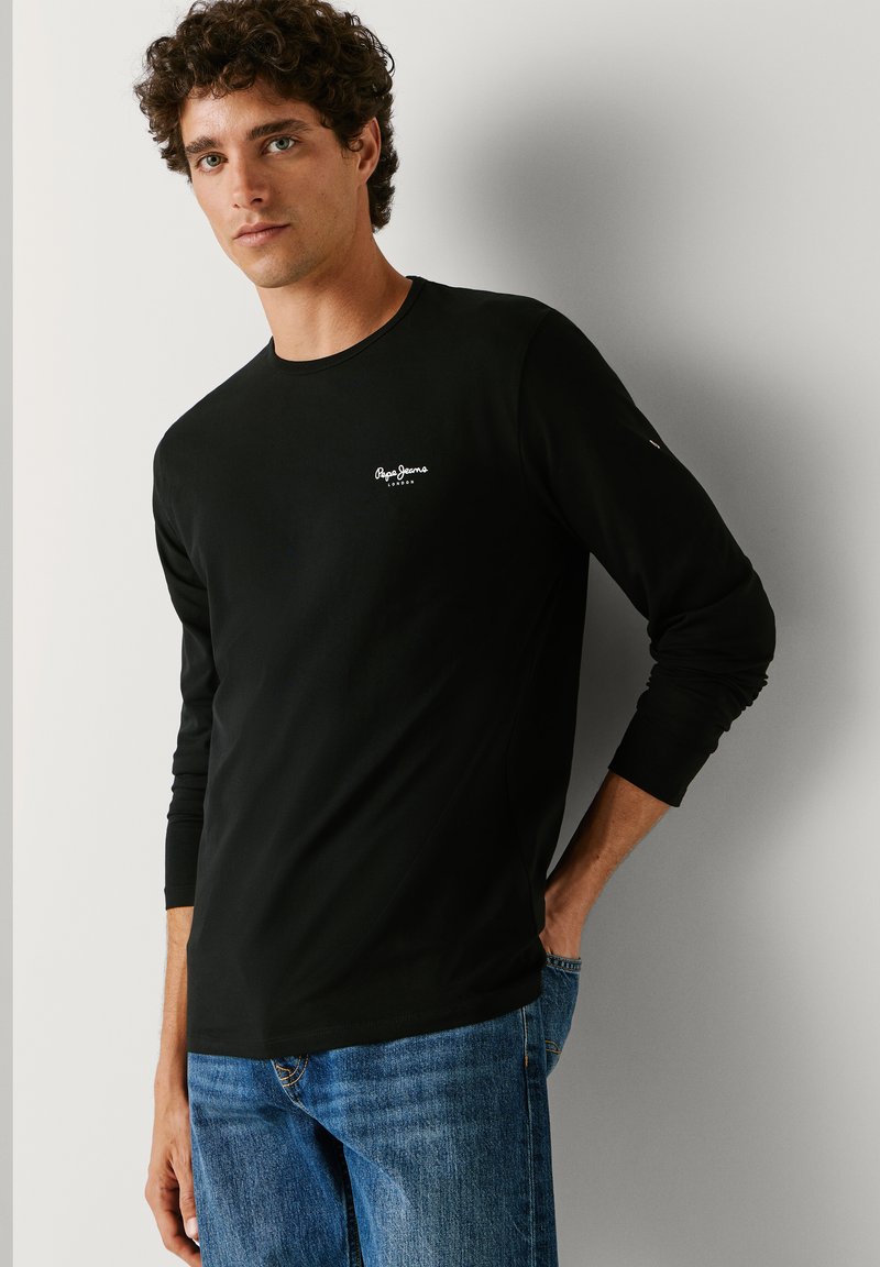 Mężczyzna z kręconymi włosami, ubrany w czarną, długorękawową koszulkę Pepe Jeans i niebieskie dżinsy, stojący z jedną ręką w kieszeni na tle białej ściany.