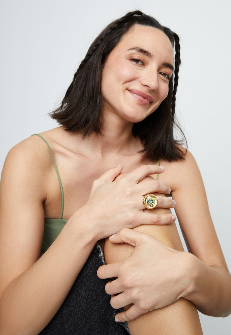 Anillo de oro con diseño de flor y una piedra preciosa azul, llevado en una mano, apoyada contra una rodilla. Se ven una prenda superior verde suave y pantalones vaqueros grises.