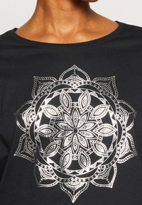 Černé tričko s velkým bílým mandalovým vzorem s propracovanými detaily, včetně kruhových a listových tvarů a dekorativních akcentů.