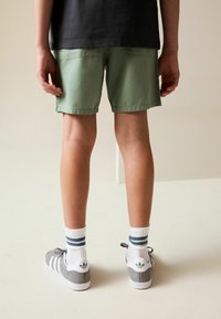 Pantaloncini verdi in cotone con tasche posteriori, abbinati a sneaker grigie con dettagli bianchi e calzini bianchi a righe. Design minimalista.