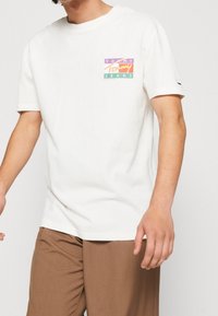 Camiseta blanca de algodón con mangas cortas que presenta un colorido logo rectangular en el lado izquierdo del pecho y un pequeño logo en la manga.