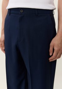 Moški, oblečen v temno mornarsko modre, po meri izdelane hlače in belo brezrokavnik, zataknjen v hlače, stoji z rokami sproščeno ob strani.