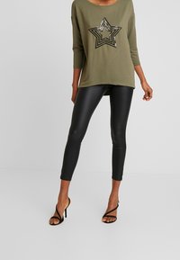 Top de manga larga verde oliva con un diseño de estrella con lentejuelas; combinado con leggings ajustados imitación piel negra y tacones negros con tiras.