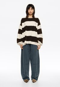 Prevelika srajca z dolgimi rokavi z bež in temno rjavimi črtami, kombinirana z ohlapnimi modrimi hlačami z detajli šivov in obuvali za natikanje.