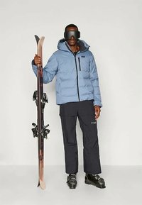 Ανοιχτό μπλε μπουφάν με φερμουάρ και λογότυπο, μαύρο παντελόνι σκι, μαύρες μπότες σκι και σκι με σταθεροποιήσεις, φορεμένα από ένα άτομο με γυαλιά σκι.