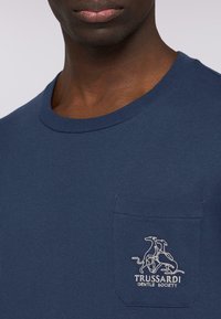 Camiseta de algodón azul marino con cuello redondo, que presenta un bolsillo en el pecho bordado con un logo que representa galgos en blanco.