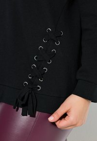 Felpa nera con dettagli incrociati in pizzo e occhielli argentati sopra l'orlo, indossata con pantaloni in pelle viola e una mano appoggiata nei paraggi.