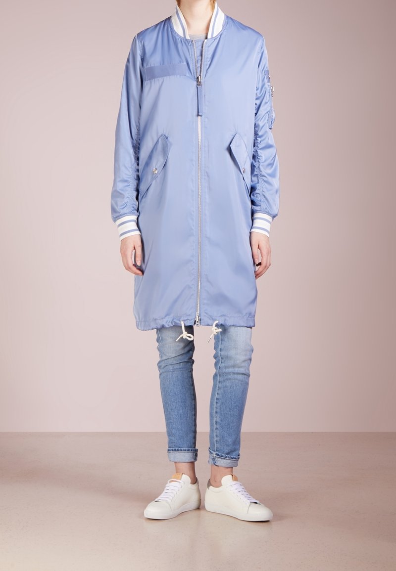 Hellblaue Bomberjacke mit weißen Rippbündchen, Reißverschluss, Seitentaschen und einstellbarem Kordelzug am Saum. Getragen über Jeans und weißen Sneakers.