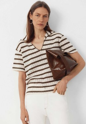 Kvinde i brun- og hvidstribet striktrøje holder stor foldet brun lædertaske, iført hvide bukser og hoops øreringe.