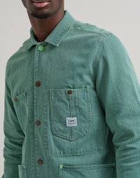 Veste en toile rayée verte avec un devant boutonné, deux poches avant et une étiquette. Présente des boutons contrastés et un tissu texturé.