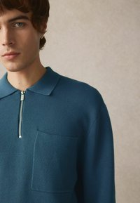 Teal striketrøje med polo, der har en halv-zip lukning, krave og en brystlomme. Strukturen er ribbet med en glat overflade.