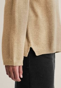 Beige stickad tröja med långa ärmar, ribbade mudd och sidospaltor; bärs över en svart underdel och visar en mjuk textur.