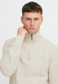 Kremasti pleteni pulover s visokim ovratnikom i zatvaranjem na zip. Ima teksturirane uzorke i rebraste detalje na rukavima i tijelu.