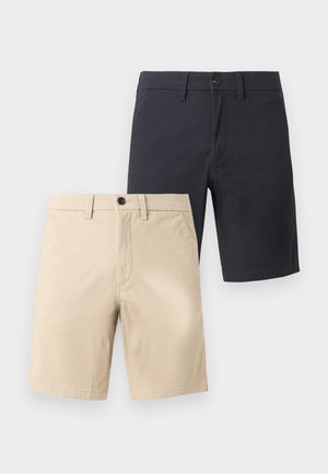 Άνδρας που στέκεται φορώντας ανοιχτό γκρι μακρυμάνικο πουκάμισο, μαύρα σορτς μέχρι το γόνατο, μαύρες κάλτσες και μαύρα loafers σε απλό φόντο.