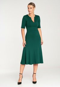 Zielona sukienka do kolan z krótkimi rękawami, dekoltem w kształcie litery V, dopasowaną górą i rozszerzanym dołem, wykonana z gładkiego materiału; noszona z czarnymi szpilkami.