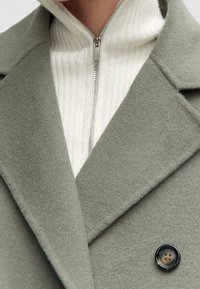 Manteau en laine vert olive avec larges revers et un seul bouton noir ; superposé sur un pull blanc côtelé à fermeture éclair. Texture douce et coupe ajustée.