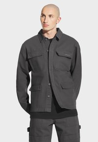 Szara kurtka zapinana na guziki, wykonana z mocnej tkaniny, z dwoma kieszeniami na piersi i luźnym krojem, w zestawie z czarną koszulą pod spodem.