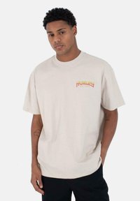Camiseta oversized de color beige claro, hecha de algodón, que presenta un logo colorido de "Hurley" en la parte izquierda del pecho. Mangas cortas y cuello redondo.