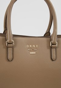 Sac à main en cuir beige avec double poignée, surface texturée, logo doré "DKNY NEW YORK" et une petite étiquette dorée "EST. 1989."