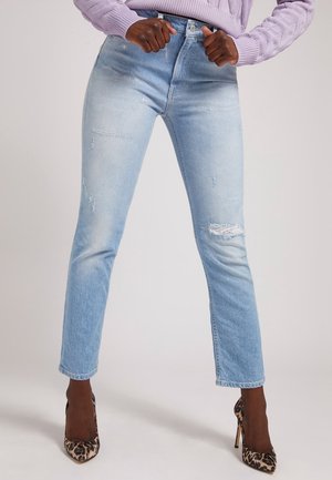 Kobieta nosząca jasnoniebieskie przetarte dżinsy, lawendowy sweter i wysokie obcasy w panterkę, stojąca z rękami na biodrach.