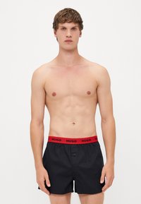 Shorts de bain noirs avec une taille élastique rouge portant la marque "HUGO", fabriqués en matériau léger, fermeture par bouton à l'avant.