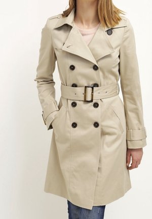 Beżowy dwurzędowy płaszcz przeciwdeszczowy z czarnymi guzikami i pasującym paskiem, noszony na jasnoróżowej bluzce i niebieskich dżinsach.