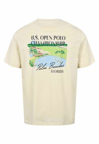 Beige T-shirt med en farverig grafik af en søbredsscene i Palm Beaches og teksten "U.S. Open Polo Championship Palm Beaches Florida" på ryggen.