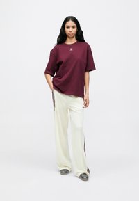 Bordó túlméretezett póló kis logóval a mellkason, krémszínű nadrággal, bordó csíkkal, és ezüst papuccsal párosítva.