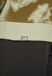Gros plan sur l'ourlet d'un sweat-shirt à motif camouflage vert avec un poignet côtelé et une petite étiquette grise du logo The North Face au-dessus d'un pantalon noir.