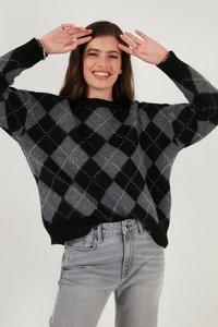 Maglione lavorato a maglia argyle nero e grigio con scollatura e polsini a coste, abbinato a jeans in denim grigio chiaro. Il modello sorride con le braccia alzate.