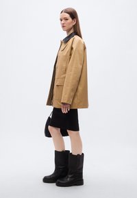 Καφέ δερμάτινο μπουφάν με μαύρο γιακά, συνδυασμένο με μαύρο εφαρμοστό φόρεμα και μαύρες μπότες μέχρι το γόνατο. Το μοντέλο κρατάει μια μαύρη υφαντή τσάντα.