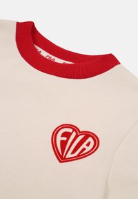 Suéter de tela crema, con cuello de canalé rojo y un logo rojo en forma de corazón que presenta "Fila" en el centro. Textura suave y diseño casual.