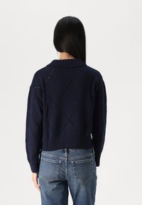 Person mit langen dunklen Haaren, der einen strukturierten navyblauen Pullover mit Rautenmuster und blaue Jeans trägt, von der Kamera weggedreht.
