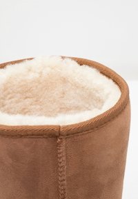 Botín de ante marrón con un suave forro de borreguito de color crema. Presenta un acento cosido en la parte superior y una textura exterior suave.