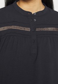 Chemise noire boutonnée avec un col mao, présentant un détail décoratif en dentelle sur la poitrine et un tissu texturé sur toute la surface.