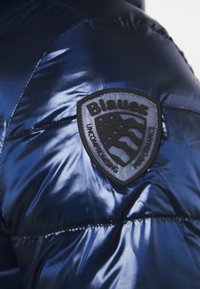 Mørkeblå pufferjakke med blank overflade. Har et sort skjoldformet mærke med teksten "Blauer" og et emblem med stjerner og striber.