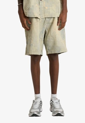 Persona con pantaloncini patchwork beige chiaro, calzini bianchi alla caviglia e scarpe da corsa bianche e grigie, in piedi su sfondo bianco.