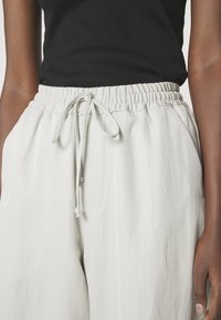 Personne portant un pantalon élastique écru avec un cordon à la taille, associé à un haut noir sans manches, debout avec une main visible.