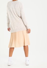 Person steht mit dem Rücken zur Kamera, trägt einen hellbeigen Pullover, einen plissierten, hellpfirsichfarbenen Rock, weiße Socken und weiße Sneaker vor einem weißen Hintergrund.