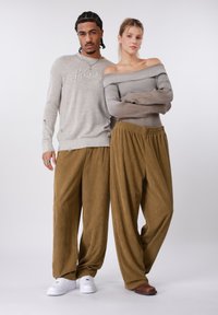 Δύο μοντέλα στέκονται το ένα δίπλα στο άλλο. Ο άνδρας φοράει ανοιχτό γκρι πουλόβερ με διακριτικές φθορές και μπεζ παντελόνι. Η γυναίκα φοράει ριμπ τοπ με ακάλυπτους ώμους και καφέ παντελόνι κορντό.