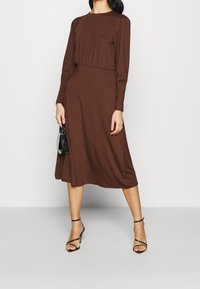 Robe midi marron avec un corsage ajusté, de longues manches bouffantes et une jupe fluide. Portée avec des sandales noires à lanières et tenant un petit sac noir.