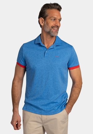 Man met baard en snor, die een blauw poloshirt met rode mouwboorden en beige broek draagt, glimlacht en kijkt naar rechts.