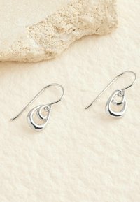 Silberne Tropfenohrringe mit drei ineinander verschachtelten Tropfenformen in polierter Optik und einem Fischhaken-Design. Texturierter beiger Hintergrund.