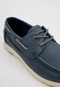Zapato de barco de cuero azul con suela blanca, que presenta cordones en contraste blanco y negro, puntera redondeada y detalles de costura en la parte superior.