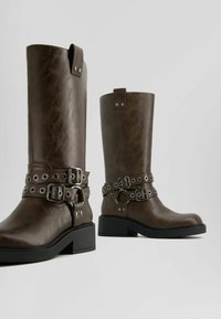 Hnědé kožené vysoké boty až po kolena s dvojitými přezkami a kovovými očky, masivním podpatkem a mírně texturovaným povrchem.