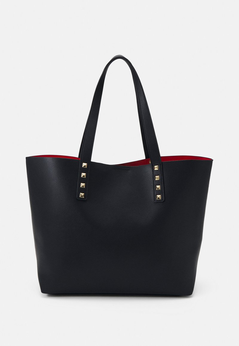 Sac fourre-tout noir avec deux poignées plates, accents en clous dorés et un intérieur rouge vif. Fabriqué en matériau lisse et texturé. Design à ouverture supérieure.