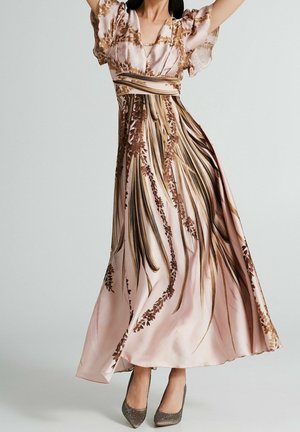 Vrouw draagt een vloeiende lichtroze maxi-jurk met bruine botanische print en vlinderachtige mouwen, gecombineerd met zilverkleurige pumps met puntige neus.