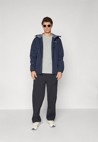 Homme portant des lunettes de soleil, une veste marine, un t-shirt gris, un pantalon cargo noir et des baskets blanches, se tenant devant un fond clair.